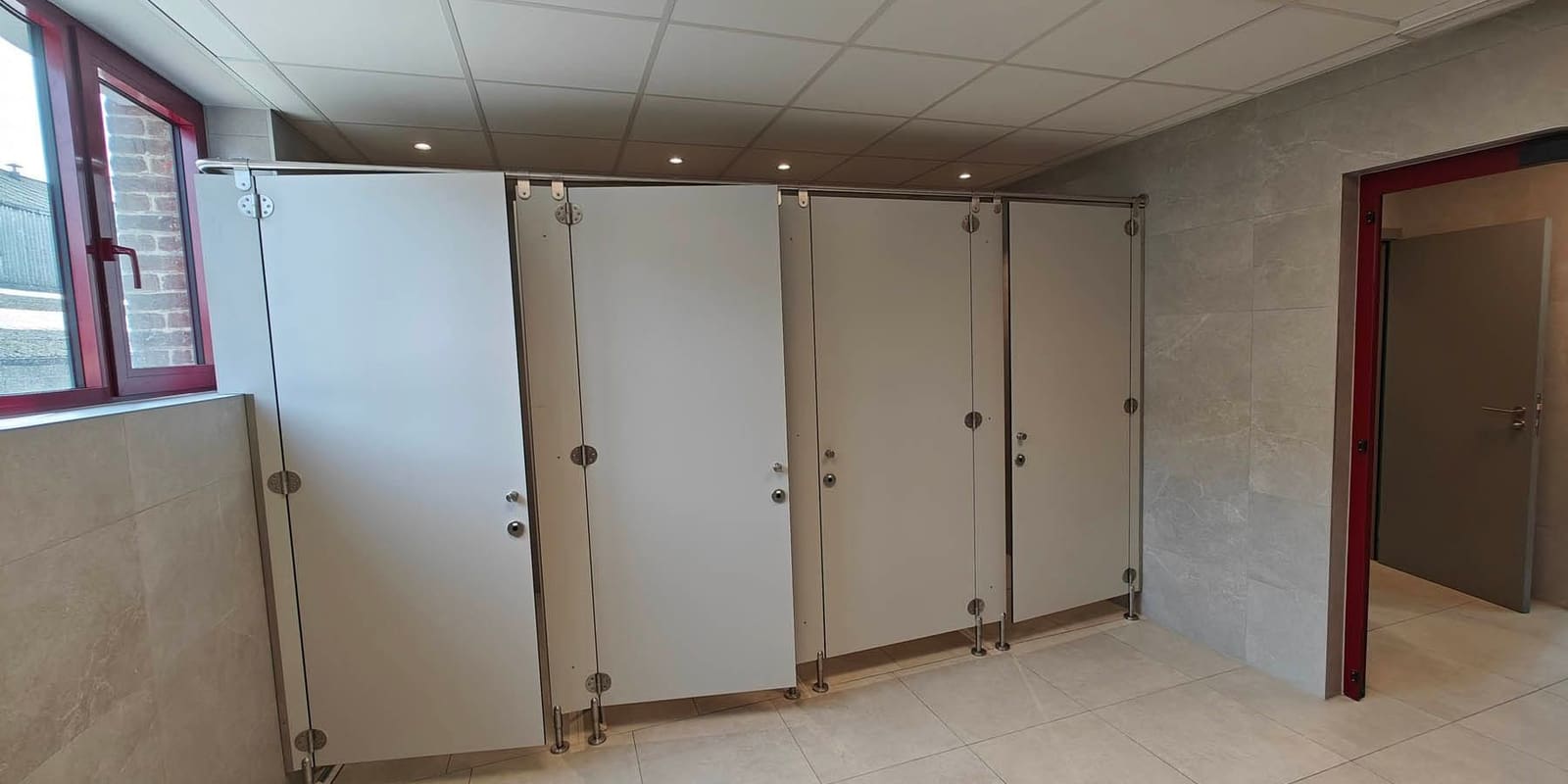 Bloc sanitaire collectif terminé : cabines en panneaux compacts sur pieds inox, carrelage grand format et faux plafond avec spots