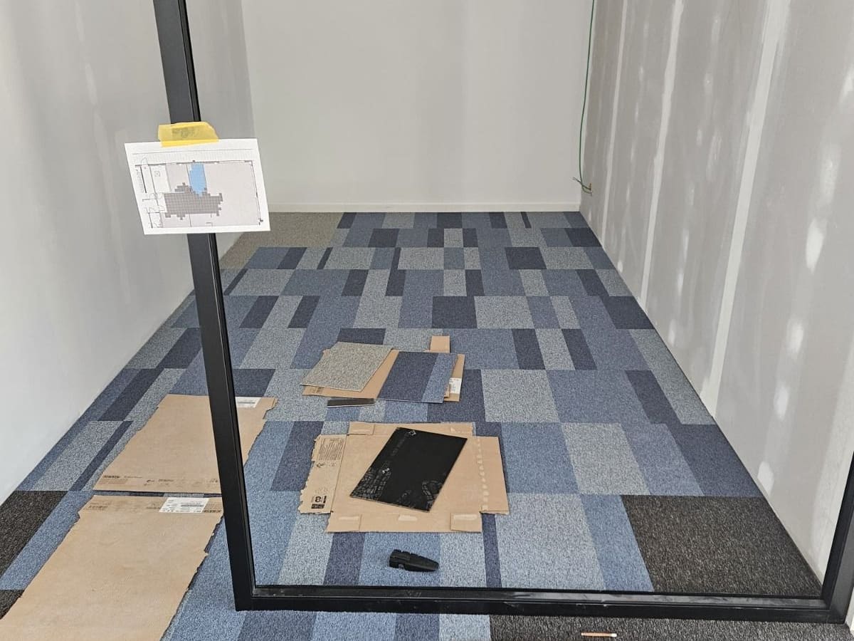 Dalles de moquette bleues posées en pose bâton rompu dans un bureau cloisonné, découpes en cours sur le pourtour