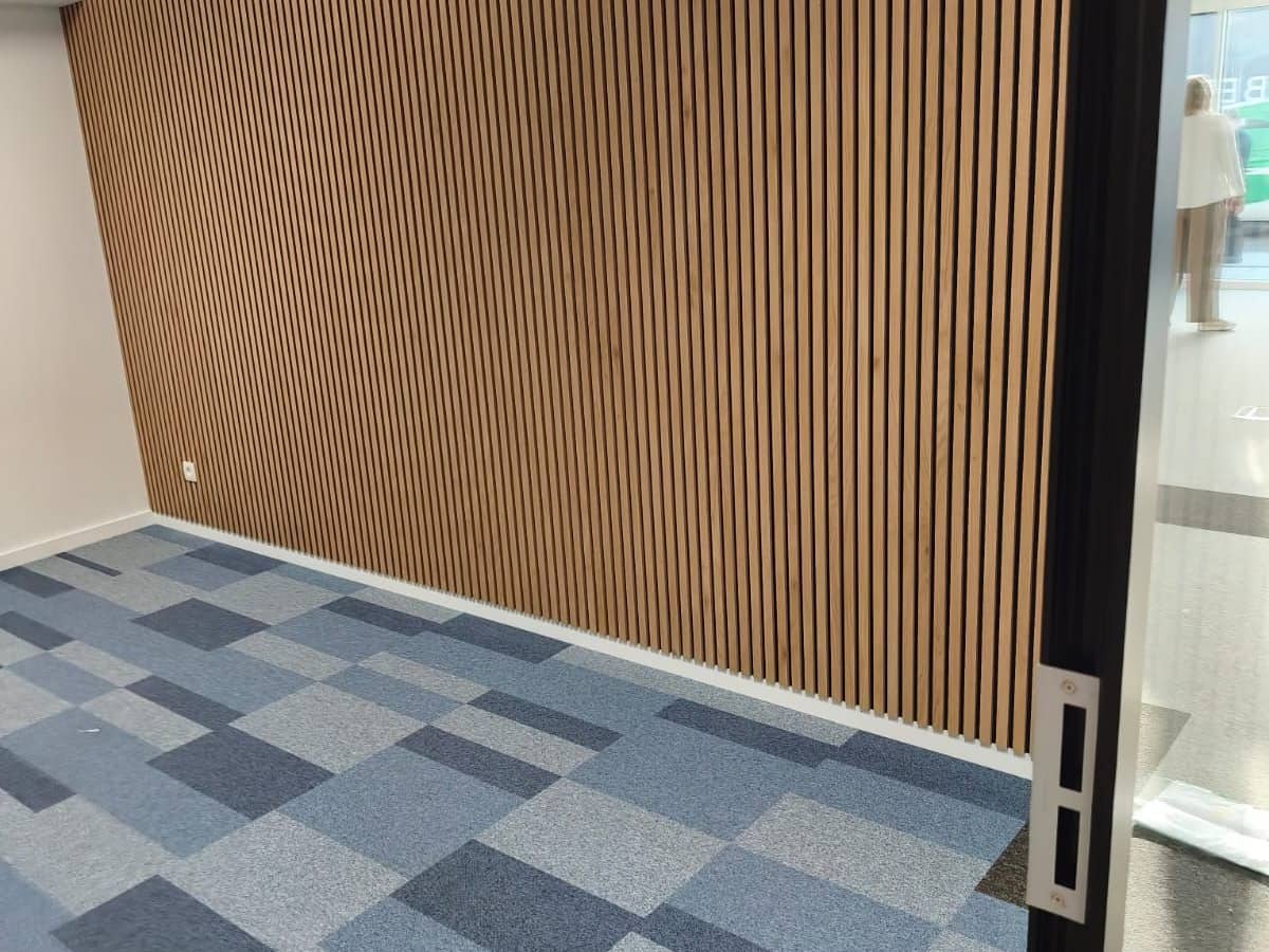 Mur habillé de tasseaux de bois verticaux sur toute sa longueur, plinthe blanche et sol en dalles de moquette bleues et grises