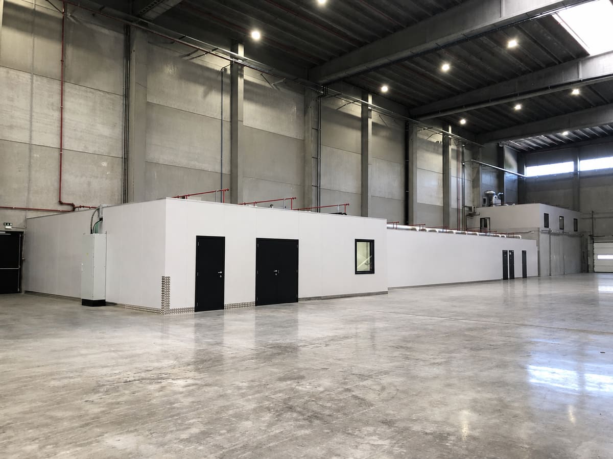 Bloc de locaux terminé à l'intérieur d'un hall industriel : parements blancs, portes métalliques noires, châssis intérieur et grilles de ventilation