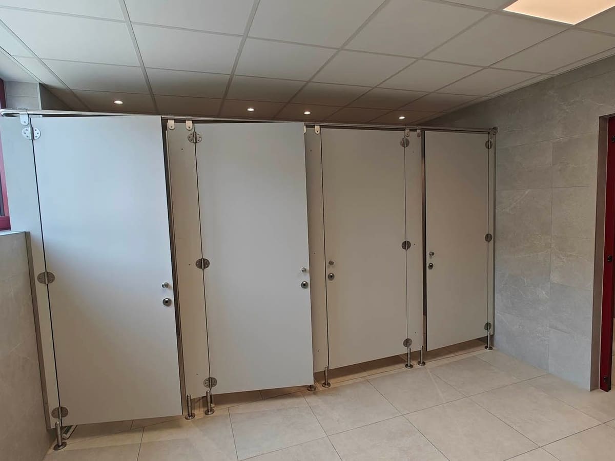 Bloc sanitaire collectif terminé : quatre cabines en panneaux compacts sur pieds inox, carrelage grand format et faux plafond avec spots