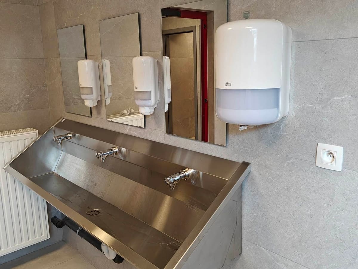 Lave-mains collectif en inox à trois postes, miroirs, distributeurs et carrelage mural grand format posé jusqu'au plafond