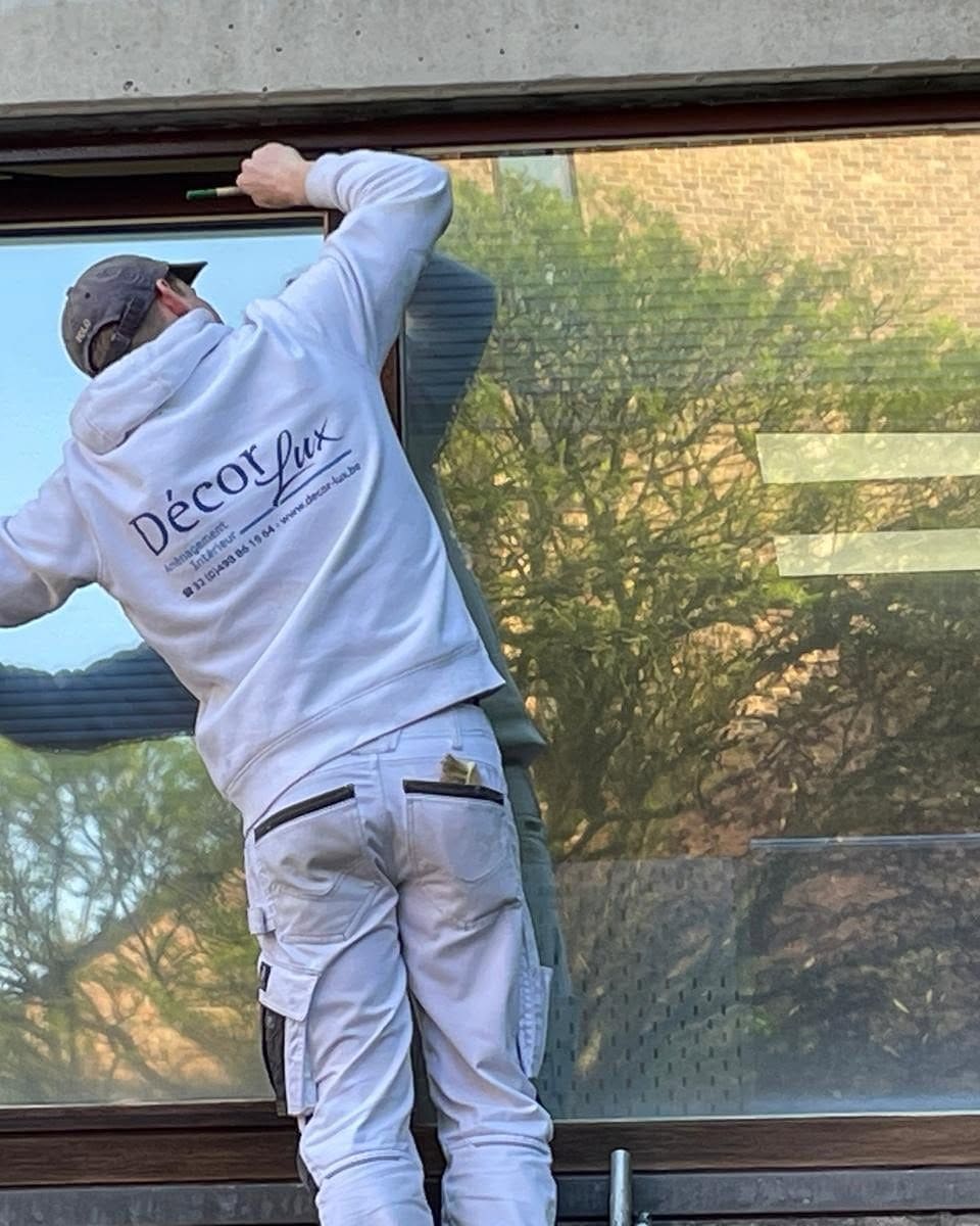 Un peintre de DECOR-LUX, en vêtements de travail marqués au nom de l'entreprise, prépare un châssis de fenêtre avant mise en peinture