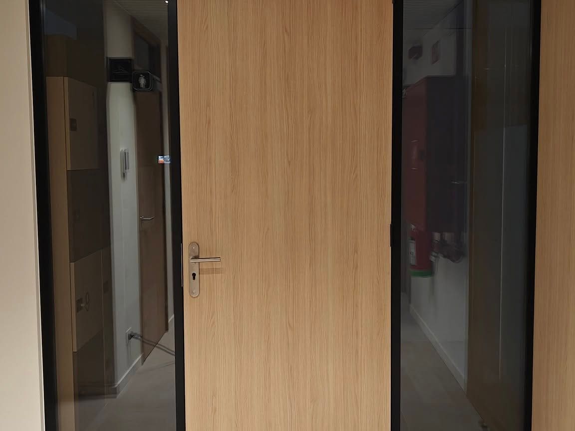 Bloc-porte stratifié bois encadré de deux châssis vitrés à profilés noirs, sous un plafond en lames de bois