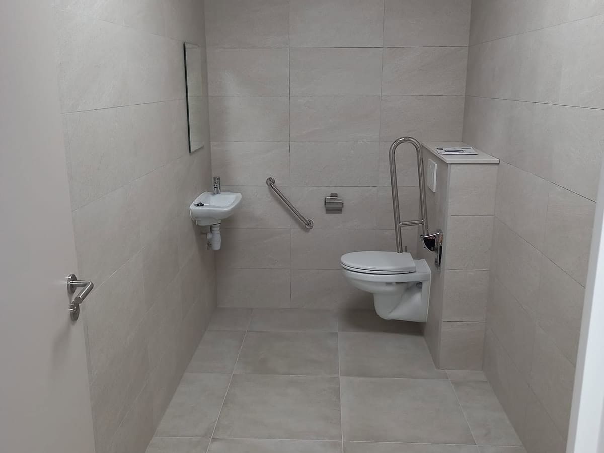 Sanitaire adapté aux personnes à mobilité réduite : WC suspendu, barre d'appui relevable, lave-mains bas et carrelage clair du sol au plafond
