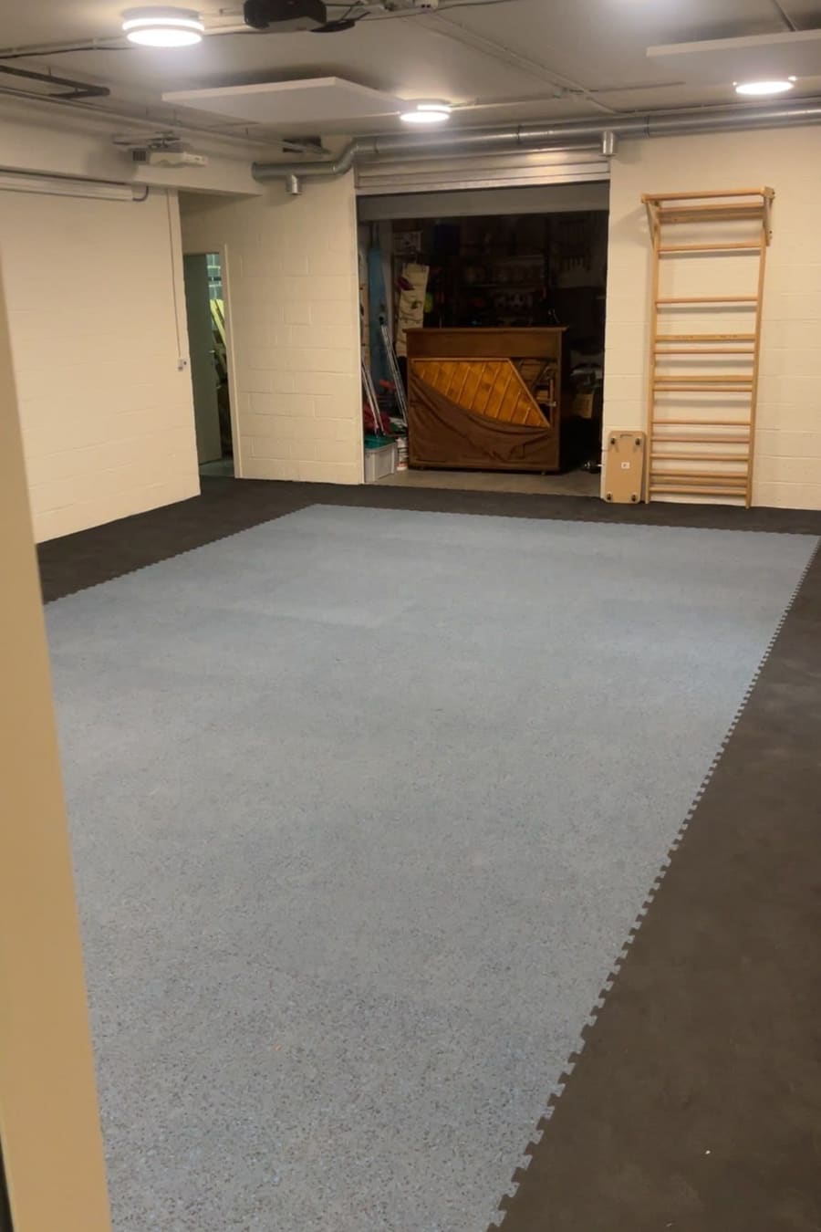 Vue d'ensemble de la salle de sport après travaux — aire d'entraînement en dalles souples gris clair bordée de dalles gris foncé, espalier mural et murs peints en blanc